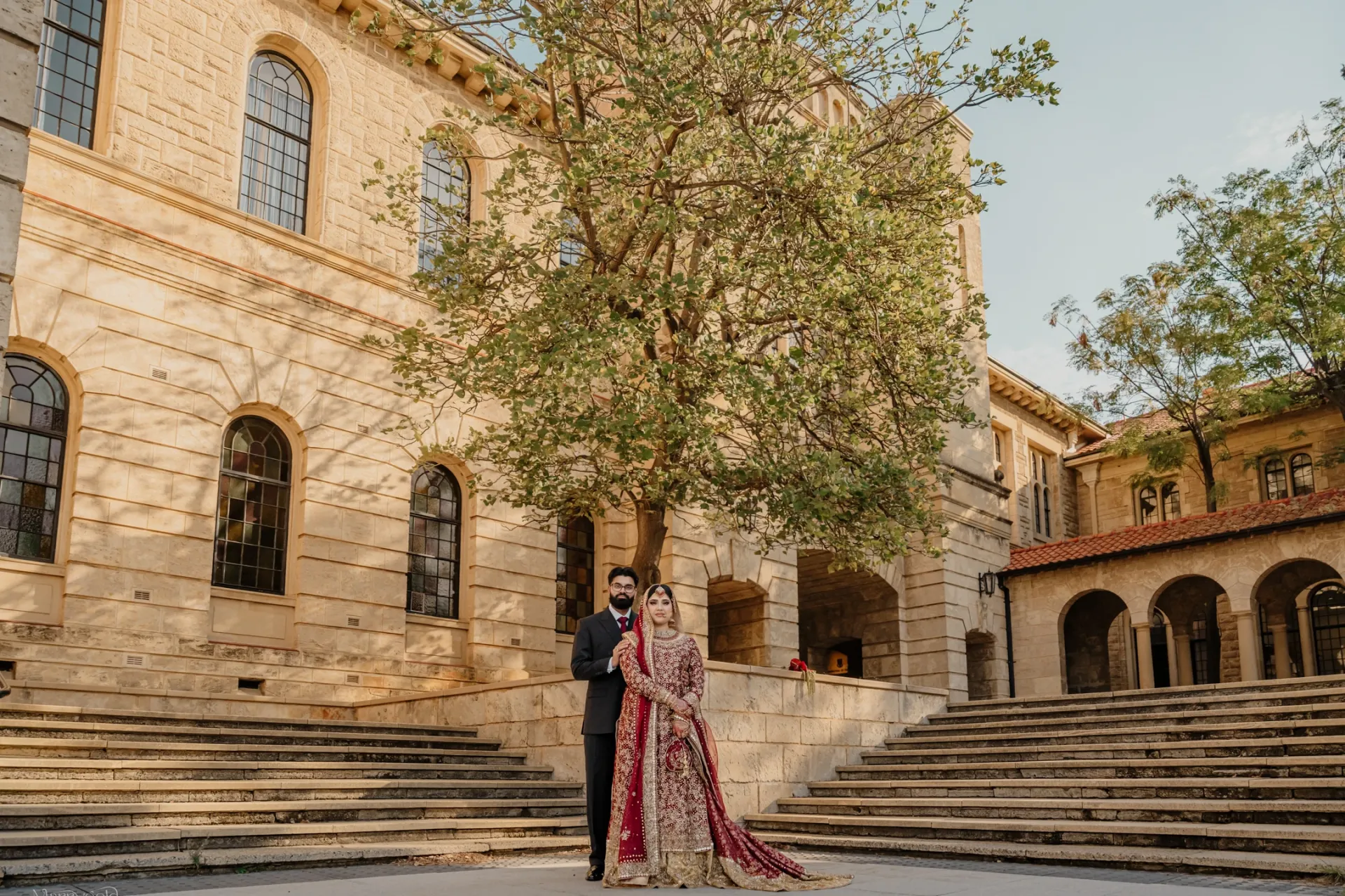 Home 22 Marrygold Weddings #1 Wedding photography and videography in Perth Groom in a dark suit and bride in a red embroidered outfit standing at the foot of a sandstone staircase beside a tree